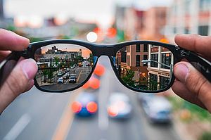 Zwei Hände halten eine Brille vor einen verschwommenen Hintergrund. Durch die Brillengläser kann man die Szenerie einer Großstadtstraße erkennen