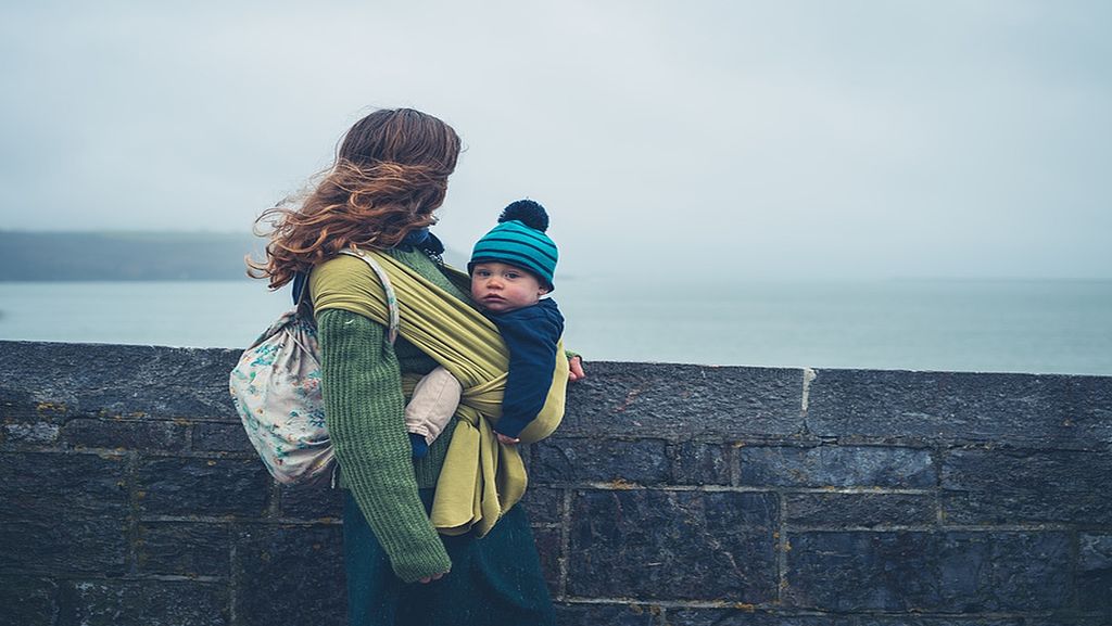 Eine Frau steht an einer Mauer und blickt an einem trüben Tag aufs Meer hinaus. In einem Tragetuch am Bauch trägt sie ein Baby, das in die Kamera blickt.