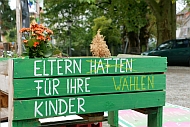 Auf einem Hochbeet aus Holz steht Eltern haften (durchgestrichen) wählen für ihre Kinder.